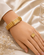 A close-up of a person's hand and wrist wearing a gold bracelet and matching gold ring, styled with a cream-colored sleeve and draped satin fabric.