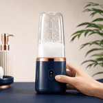 A handheld electric blender with a navy blue base, rose gold collar, and clear cup, being held by a hand on a dark blue table. The background is a softly blurred beige wall with a green plant.