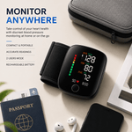 A wrist blood pressure monitor with a black strap and round digital display, shown on a light beige tabletop with a gray zip case, passport, earbuds, and a smartphone nearby. Text on the left reads 'MONITOR ANYWHERE' with feature callouts.